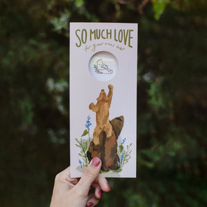 No. 10 size greeting card that includes a suncatcher sticker inside. Front: Baby bear standing on mama bear's back with words "So much love for your new cub." Colors are warm tones of pinks, browns, and greens.